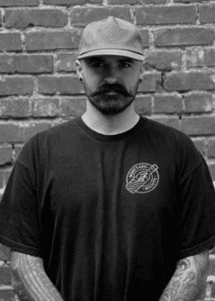 A man with a beard and cap is standing in front of a brick wall.
