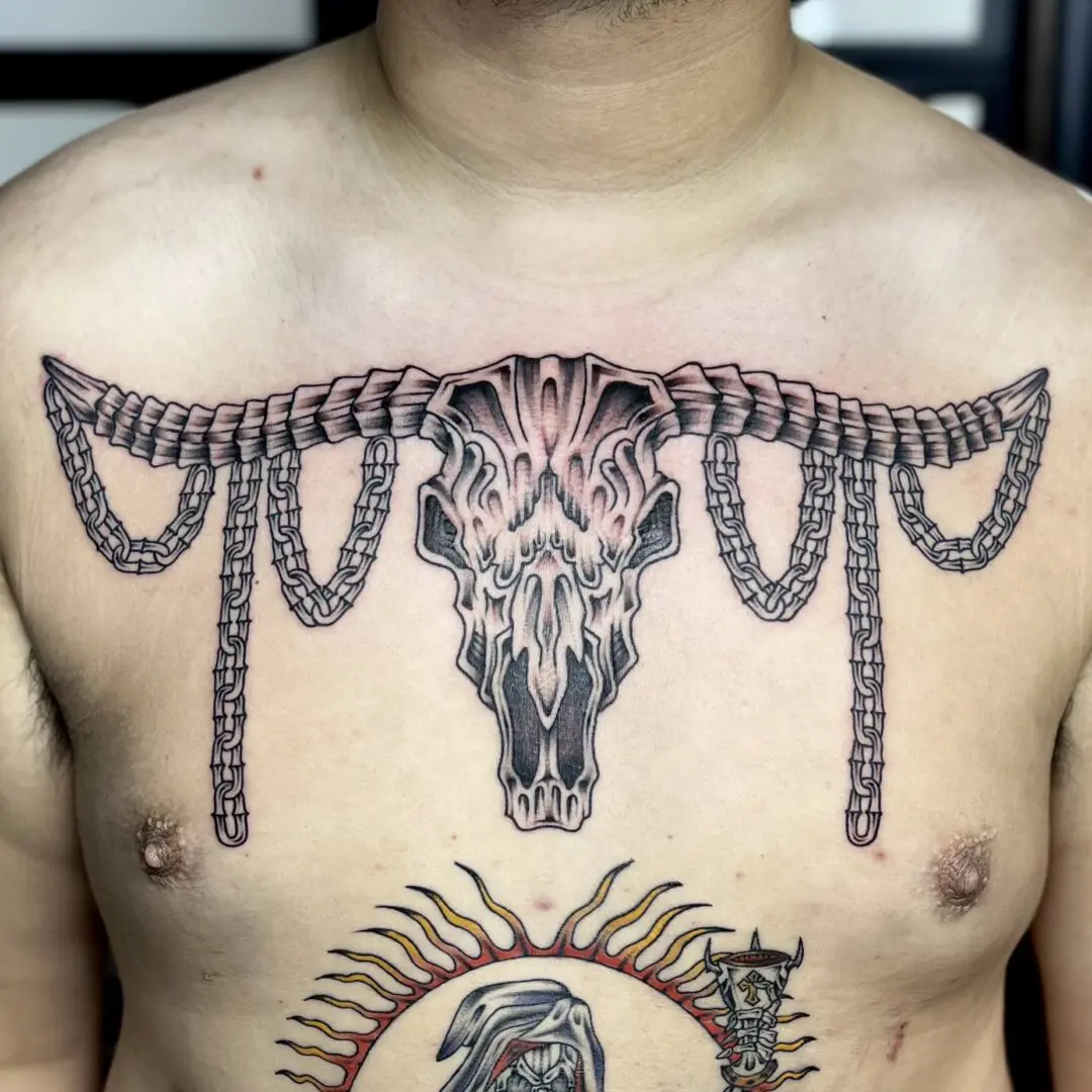 A man with a black and gray traditional tattoo of a cow skull on his chest.